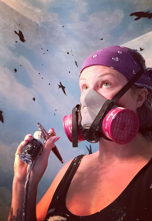 artist Jennifer Little holds an airbrush gun in room with airbrushed sky and birds
