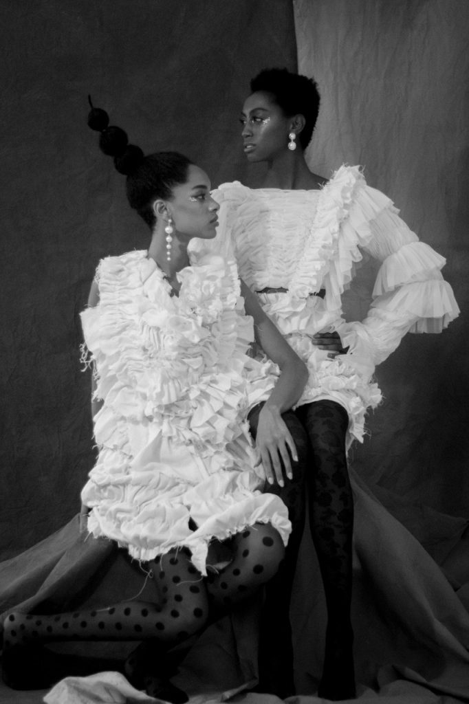 two black models in white, ruffled dresses sitting on stool, makeup and hair assist by Indonesia's Melfinna Tjugito