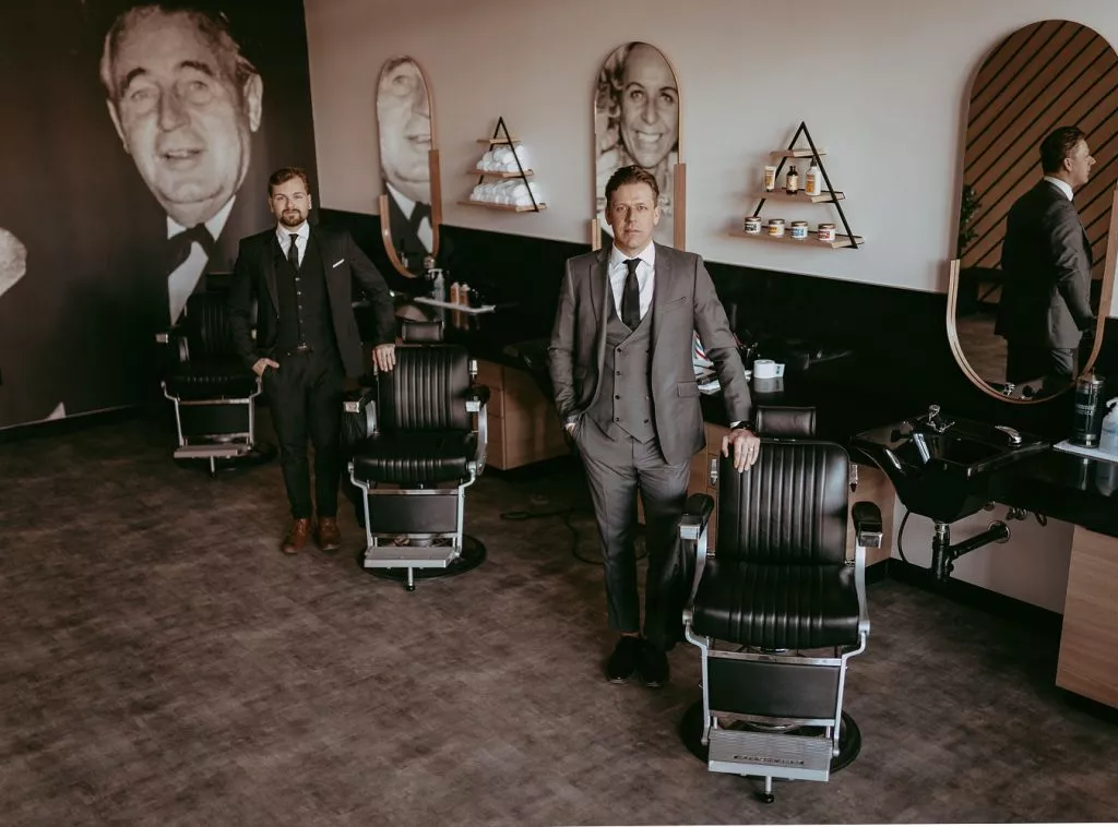 Blanche Macdonald Graduates Cody Dunbar and Jason Hatheway pose in suits, leaning against barbering chairs inside of Black Forest Barbershop