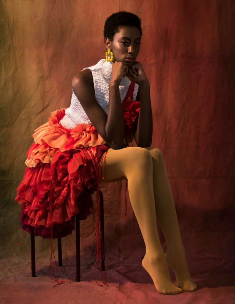 Model sits on stool wearing nude pantyhose and layered orange skirt, artist Melfinna Tjugito