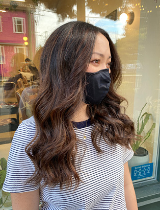 a women wearing a black mask, standing in front of Stone Fox Salon, with long-length soft curls hairstyles in brown, styled by Kevin Murphy Colour Educator Kalli Wyssen