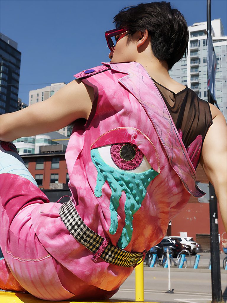 a male model with a tie-dyed canvas one-shoulder vest with side zip, asymmetrical lapel with a black micro mesh tank top.