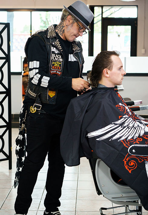 Ash, wearing a grey fedoras hat, trimming a customer's back hair 