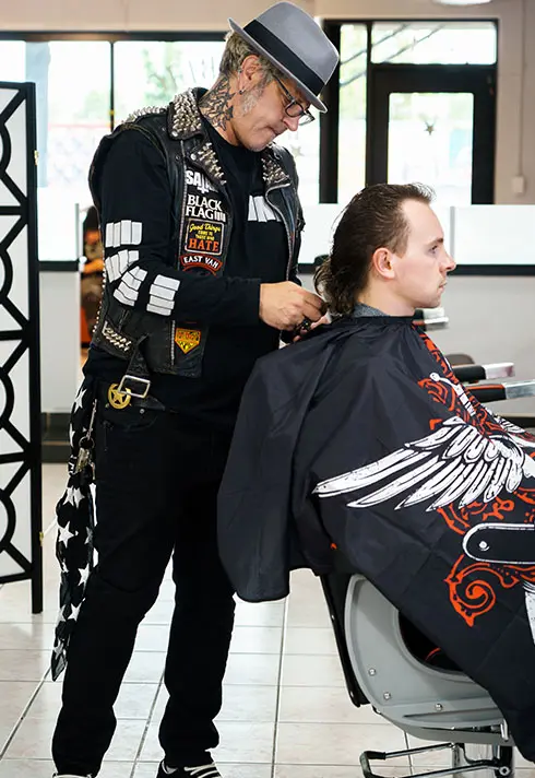 Ash, wearing a grey fedoras hat, trimming a customer's back hair