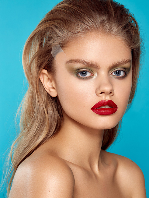 A Causian female model wearing a bright red lip stain with light green eyeshadows posing infront a teal blue backdrop.